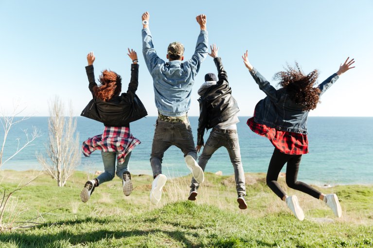 Back view picture of a group of friends jumping outdoors near beach with raised hands.